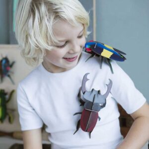 A blond child wearing a white T-shirt smiles as they look down at two large, colourful Rosalia Beetle toys—one on their shoulder and another on their chest. Gate21.ee
