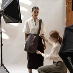 A woman poses in front of a white backdrop holding a Feel Good Bag - Chicory Coffee, dressed in a two-tone outfit, while another woman adjusts her clothing. Studio lights and equipment are visible around them in the photo studio. Gate21.ee