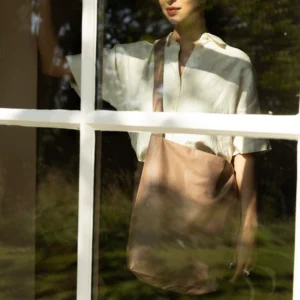 A person stands behind a window, looking outside, wearing a light-coloured shirt and carrying a tan Feel Good Bag - Beaver Fur. Sunlight and tree shadows fall on them and the window. Gate21.ee