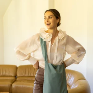 A woman stands indoors in front of a tan leather sofa, smiling and looking to her left. She wears a sheer white blouse, brown trousers, and carries the Feel Good Bag - Chinois Green by Tinne+mia crossbody, with her hands on her hips. Gate21.ee