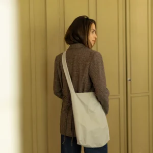 A person with shoulder-length brown hair, in a brown checked blazer and blue jeans, stands indoors near beige panelled doors, facing away from the camera with a white Feel Good Bag - Feather Grey by Tinne+mia on their shoulder. Gate21.ee