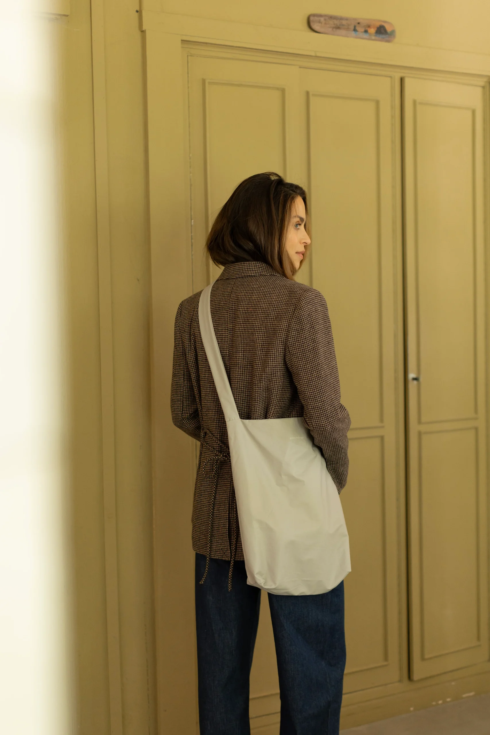 A person with shoulder-length brown hair, in a brown checked blazer and blue jeans, stands indoors near beige panelled doors, facing away from the camera with a white Feel Good Bag - Feather Grey by Tinne+mia on their shoulder. Gate21.ee