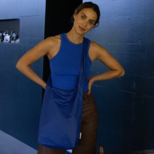A woman in a blue sleeveless top, brown trousers, and a Feel Good Bag - Sodalite Blue stands indoors with her hands on her hips in front of a dark wall and black doorway. Gate21.ee