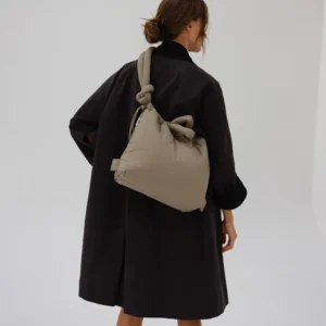 A person with brown hair in a low, messy bun wears a black coat and carries the Ona Soft Bag in Sage—its knotted straps and spacious size make it ideal for Tallinn shopping or picking up finds from an Estonian gift shop—against a plain light background. Gate21.ee