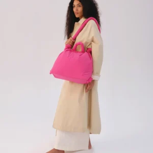 A person with long curly hair stands against a plain background, wearing a long beige outfit and carrying the Ona Soft Bag - Pink—an effortlessly curated look, perfect for shopping in Tallinn or visiting a concept shop. Gate21.ee