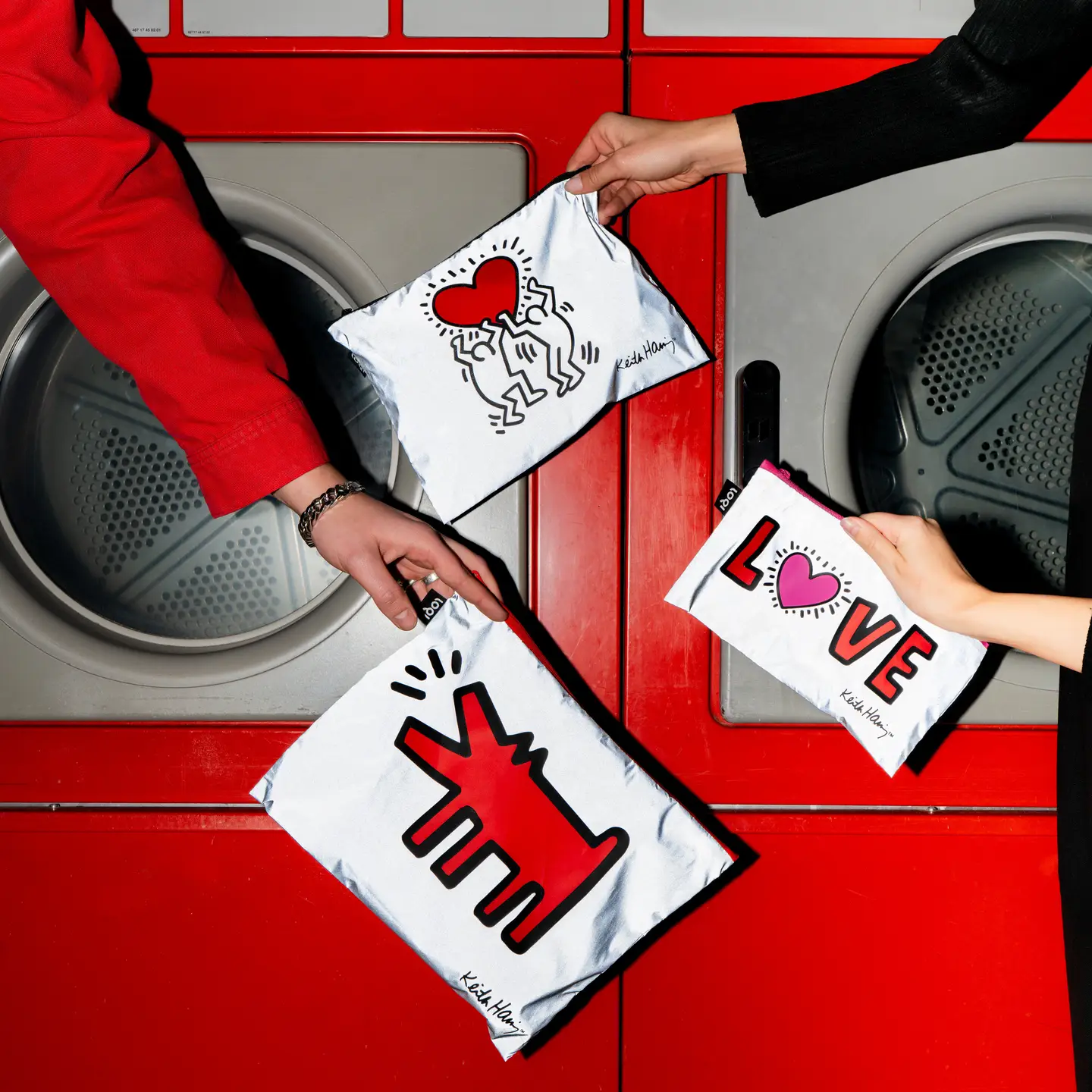 Two hands hold the Keith Haring 3x Zip Pockets LOQI Reflective pouches, each with a reflective zip pocket design—one showcases a red barking dog, another displays a radiating red heart, and the third spells LOVE with a heart as the O—in front of red washing machines. Gate21.ee