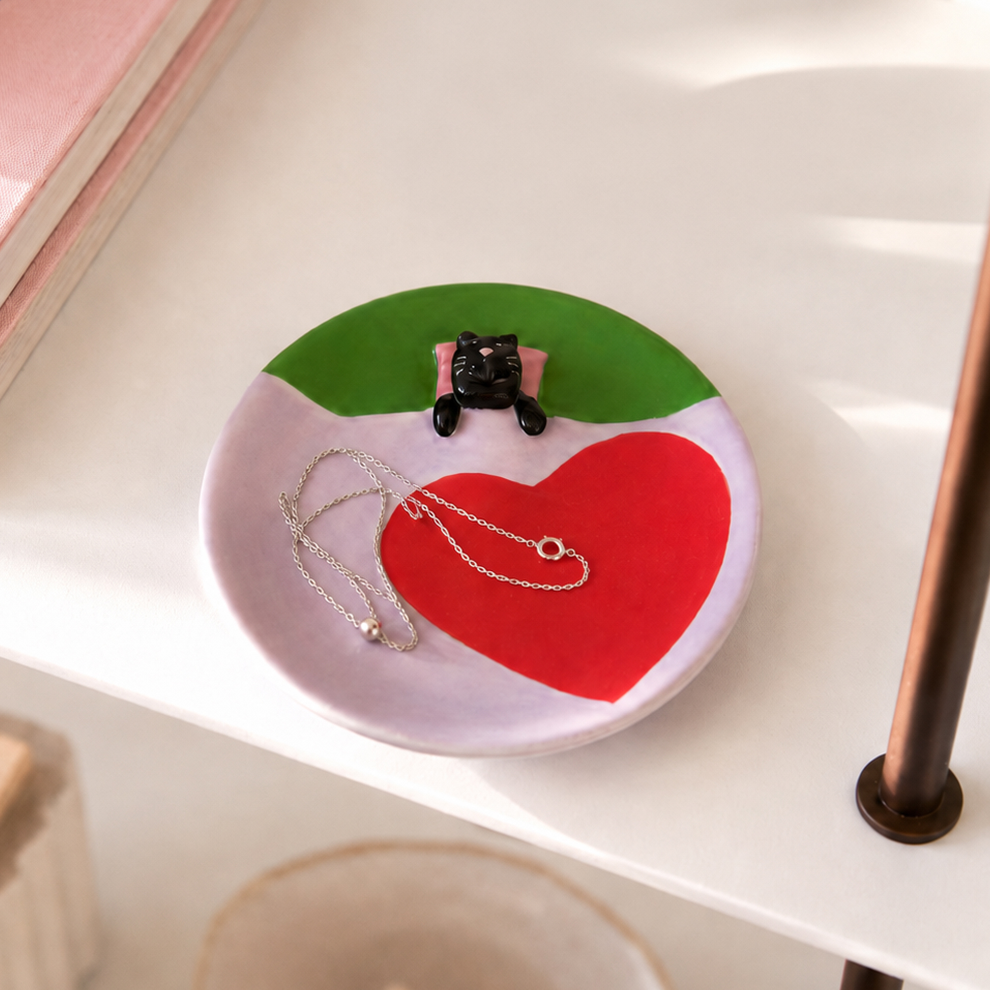A round, dreamy Pink Dreamer Cat - Jewellery Plate featuring a red heart, a green border, a small black cat figurine on a pink cushion, and a silver chain necklace resting on top. Gate21.ee