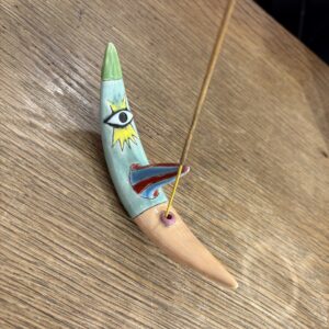 The KAURI KALLAS - Moon Incense - KÕIK holder, a crescent-shaped ceramic with painted eye and lightning bolt designs, holds a burning incense stick on a wooden surface. Gate21.ee