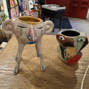 Two KAURI KALLAS - Tealight & Shots - KÕIK vases sit on a wooden surface: the left one features a face with large ears, closed eyes, and three legs; the right is angular with abstract geometric facial details in red, black, and beige. Gate21.ee