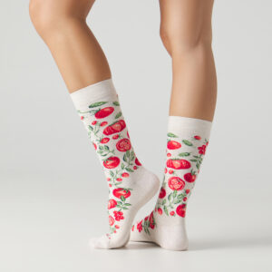 A person wearing beige Jimmy Lion Tomato Fever socks with a red tomato and green leaves pattern stands on tiptoe against a plain light background. Gate21.ee