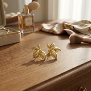 A pair of Fleur - BØLD Earrings, crafted in a gold leaf design, rest on a wooden dressing table beside a make-up brush, scarf, and perfume bottle. Gate21.ee
