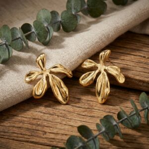 A pair of Nalani - BØLD gold-coloured, abstract flower-shaped earrings are displayed on a wooden surface beside a beige cloth and green eucalyptus leaves. Gate21.ee