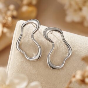 The Aversa - BØLD Earrings in silver are showcased on a light beige textured surface, with soft-focus beige and cream floral accents in the background. Gate21.ee