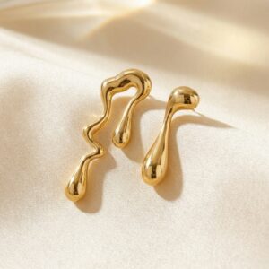 The Drops - BØLD Earrings - Gold feature two abstract, gold-toned metal pieces with smooth, fluid shapes displayed on a light cream satin fabric background. Gate21.ee