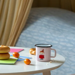 A pink Little MY - Moomin enamel mug (3.7dl) sits on a round table next to bagels, kumquats, and colourful notepads. In the background, there's a bed with a blue patterned blanket. Gate21.ee