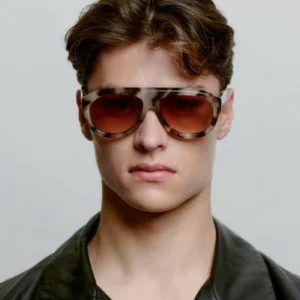 A young person with short, wavy brown hair wears large Rio - Demi Tortoise sunglasses and a black leather jacket, staring directly at the camera against a neutral background. Gate21.ee