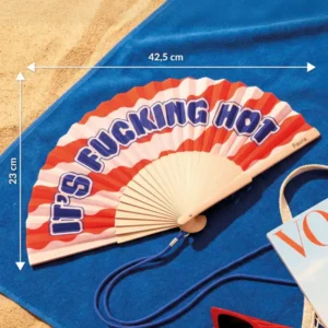 The "It's fucking hot!" - fan - Red waves, featuring bold red and pink with printed text, is open on a blue beach towel. Perfect for summer, it measures 42.5 cm wide by 23 cm tall. Gate21.ee