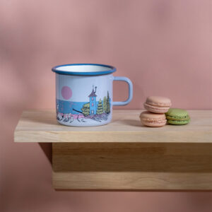 The Sunset - Moomin enamel mug 550ml in light blue with an illustrated scene sits on a wooden shelf beside three stacked macarons—green, pink, and beige—set against a pink background. Gate21.ee