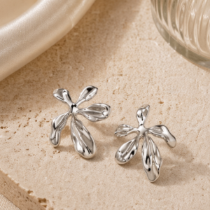 The Nalani - BØLD Earrings (Copy), shiny and abstract with a flower petal design, are displayed on textured beige stone, set against a backdrop of glass and satin fabric. Gate21.ee