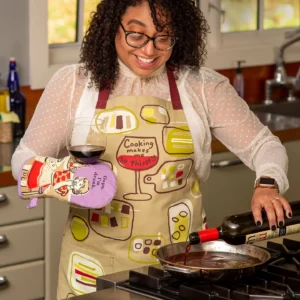 A woman in glasses and an apron smiles in her kitchen as she pours red wine from a bottle into a pan, holding a wine glass and wearing the colourful "Oops, I'm drunk" Oven Mitt by BlueQ. Gate21.ee