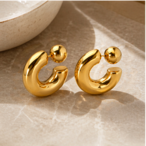 A pair of Luna Plate - BØLD Earrings - Gold with shiny, crescent shapes and spherical studs rest on a beige, textured surface by the curved edge of a light-coloured bowl. Gate21.ee