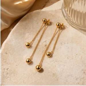 A pair of Luna Plate - BØLD Earrings - Gold features dual gold chains on each earring, each ending with a gleaming gold sphere. The earrings are shown on beige stone beside pleated fabric and a ribbed glass object. Gate21.ee
