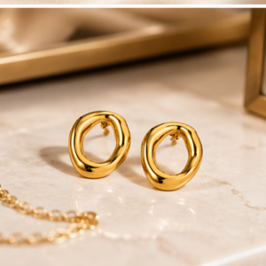 A pair of Drops - BØLD Earrings - Gold (Copy), featuring irregularly shaped circular studs, sits on a marble surface beside part of a gold chain and a glass container with a gold frame. Gate21.ee