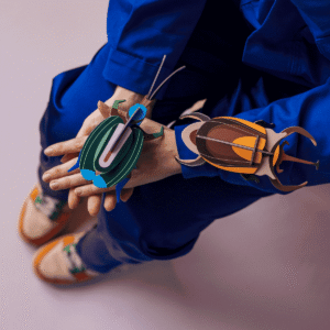 Wearing blue, a person sits with hands crossed, displaying large, colourful Stag Beetle (Copy) paper models by StudioROOF on their arm and hand. Their shoes are white with orange and green accents against a light purple background. Gate21.ee