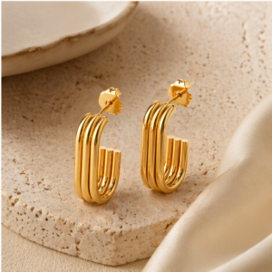 The Luna Plate - BØLD Earrings - Gold feature rectangular, gold hoops with a layered tubular design, displayed on a textured beige stone beside cream fabric and a ceramic plate. Gate21.ee