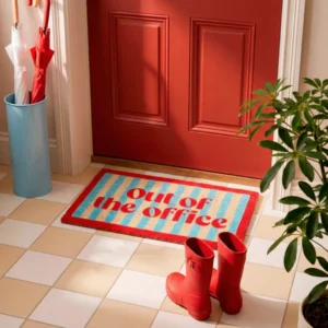 A red door with the "Out Of The Office" doormat in red letters on blue and beige stripes, nearby red wellies, a green plant, a blue stand holding red and white umbrellas, and a handheld fan for hot summer days. Gate21.ee