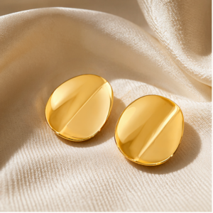 Two shiny, oval-shaped Luna Plate - BØLD Earrings in gold are displayed on cream satin fabric, reflecting light and showcasing their smooth, polished surface. Gate21.ee