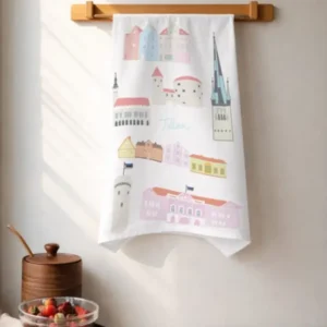 A Köögirätik – Vanalinn white kitchen tea towel with colourful building illustrations hangs on a wooden rack above a worktop with fruit, utensils, and a wooden container. A New Rebels small rucksack is nearby as soft sunlight streams in from the left. Gate21.ee