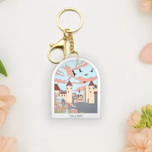 A Võtmehoidja – Talvine mets keyring with a gold clasp rests on a white surface, surrounded by peach flowers, heart-shaped objects, and a New Rebels small rucksack. The keyring features an illustrated winter forest scene. Gate21.ee