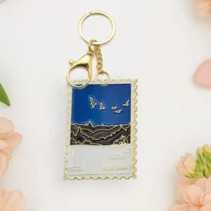 The Võtmehoidja – Talvine mets keyring, with a gold border and winter forest scene, is placed on a white surface next to pink flowers, heart-shaped items, and a chic New Rebels small rucksack. Gate21.ee