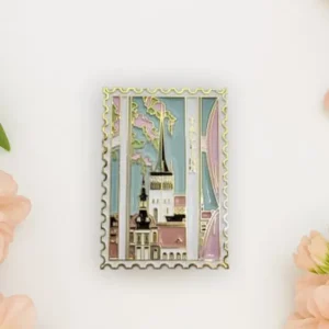 A rectangular “Magnet – Tallinna vanalinn” shaped like a postage stamp features a cityscape with church spires, displayed on a white surface with pink flowers, heart-shaped items, and a chic New Rebels small rucksack nearby. Gate21.ee