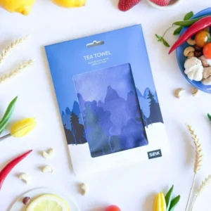 A packaged Köögirätik – Sini-must-valge lies on a white surface with lemons, strawberries, chilli peppers, sliced lemon, a yellow tulip, a blue bowl of nuts, and a New Rebels small rucksack adding style to the vibrant scene. Gate21.ee