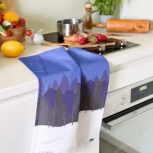 A Köögirätik – Sini-must-valge tea towel with a blue and white Estonia landscape hangs on the oven handle. On the worktop are fruit, herbs, a saucepan, and a chopping board by a New Rebels small rucksack ready for adventure. Gate21.ee