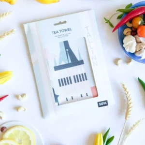 A packaged Köögirätik – Vaade Tallinnale tea towel is displayed on a white surface with lemons, strawberries, tomatoes, peppers, mushrooms, grains, and a New Rebels small rucksack arranged around it. Gate21.ee