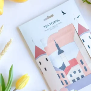 A packaged Köögirätik – Viru väravad tea towel displays buildings with red roofs, a church steeple, lemons, a strawberry, a yellow tulip, green leaves, cashew nuts, and a stylish New Rebels small rucksack on a white surface. Gate21.ee