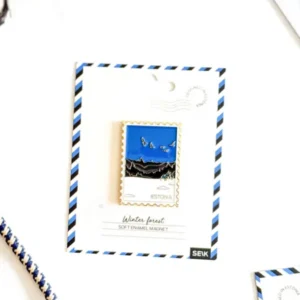 The Magnet – wintry forest, a rectangular enamel magnet featuring birds over a winter forest with "Estonia," is displayed on a white card labelled Winter Forest, set against a white background beside a striped rope and small New Rebels rucksack. Gate21.ee