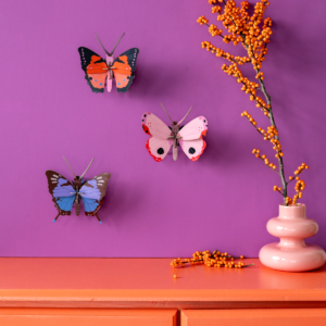 A pink vase with orange berries sits on an orange chest of drawers. Above it, three Fern Butterfly (Copy) butterflies by StudioROOF are mounted on a purple wall, adding a whimsical touch to the scene. Gate21.ee