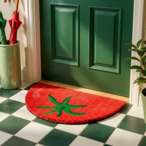 The "Half Moon Tomato" doormat, shaped like a semicircle and featuring a red tomato with a green stem, is placed by a green door on chequered tiles. An umbrella stand, potted plant, and handheld fan finish the inviting entrance. Gate21.ee
