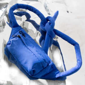 A red Knotflex Rucksack with long, padded straps tied in a loose knot and a front zip pocket is displayed on a reflective silver surface. Gate21.ee