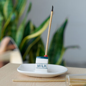 A lit incense stick rests in the "Incense burner - Milk," a modern ceramic holder shaped like a milk carton, set on a white dish atop wood, with green plants softly blurred in the background. Gate21.ee