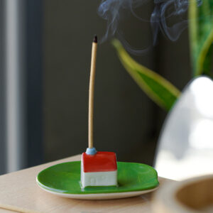 A lit incense stick stands upright in the Incense burner - Milk, a small ceramic holder shaped like a house with a red roof on a green dish. Thin smoke rises as a plant sits in the background, showcasing Helio Ferreti’s modern, colourful design. Gate21.ee
