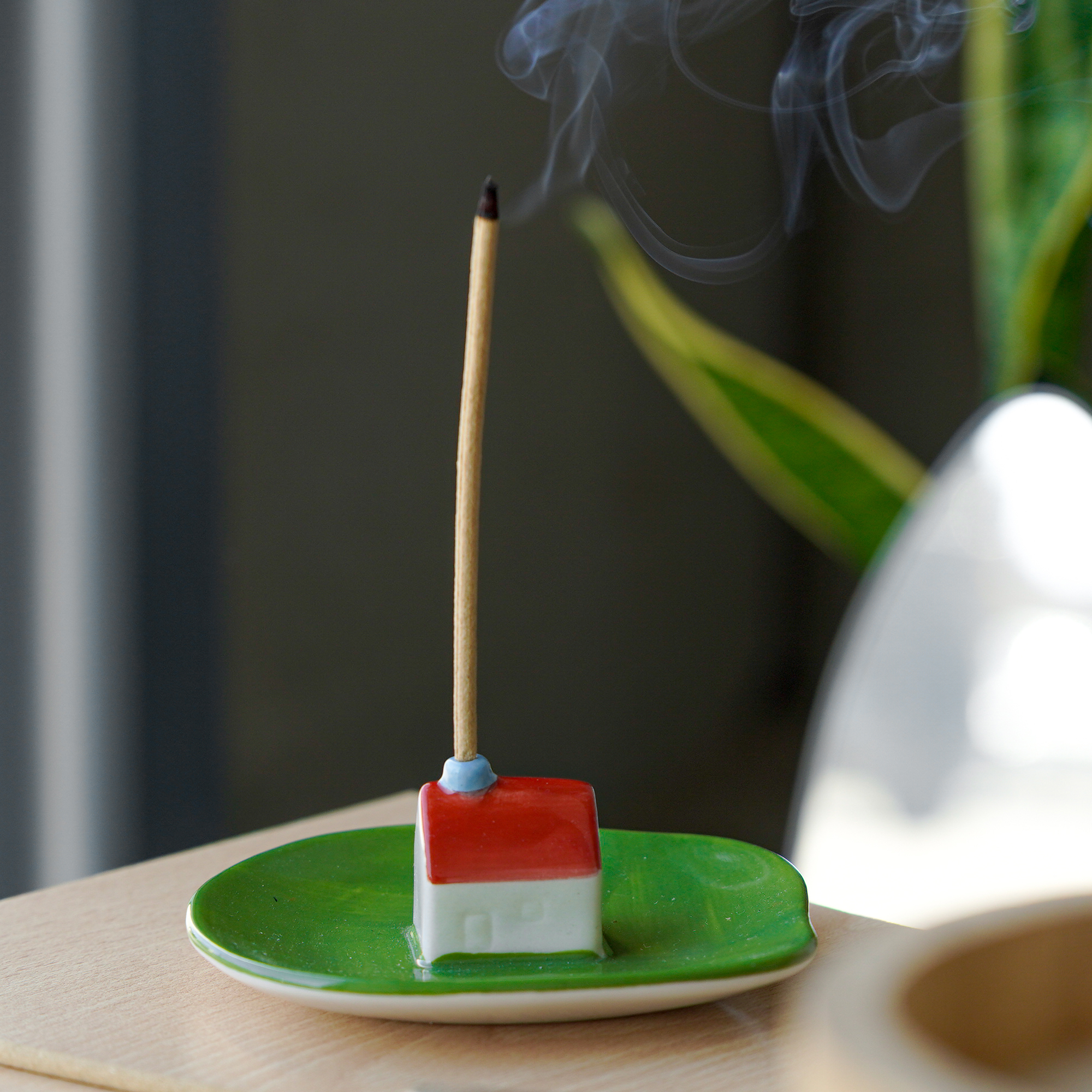 A lit incense stick stands upright in the Incense burner - Milk, a small ceramic holder shaped like a house with a red roof on a green dish. Thin smoke rises as a plant sits in the background, showcasing Helio Ferreti’s modern, colourful design. Gate21.ee