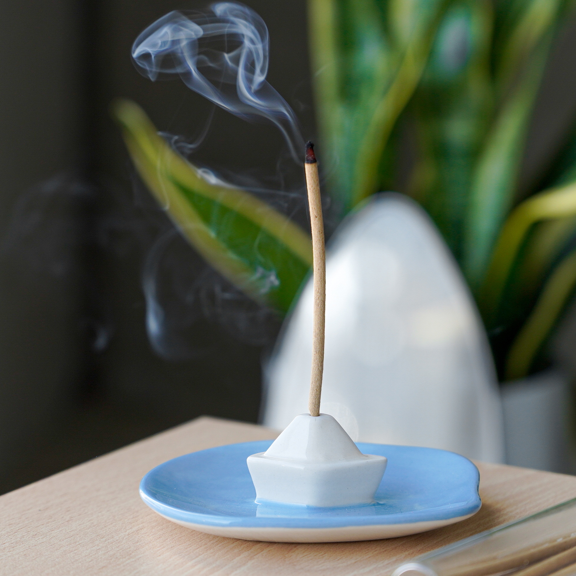 A lit incense stick stands upright in the white, boat-shaped "Incense burner - Milk" by Helio Ferreti on a blue plate, with a green plant and blurred background elements visible. Gate21.ee
