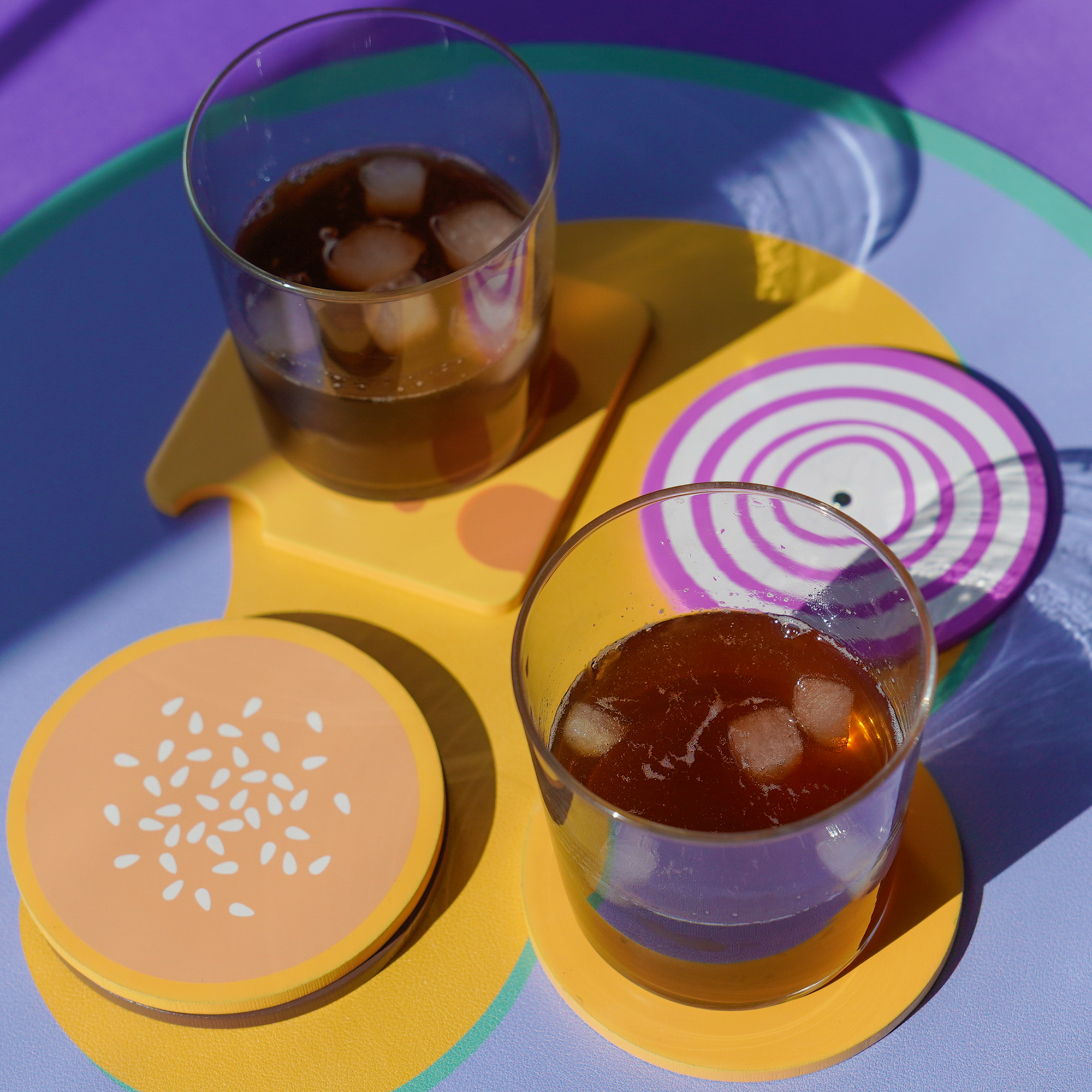 Two glasses of iced coffee sit on a colourful tray with yellow and purple coasters from the Burger - Coaster Set, adding a playful touch to the vibrant scene. Gate21.ee