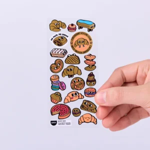 A hand holds a sheet of Croissant - Earring Stickers (Copy), featuring colourful cartoon bread and pastry characters like croissants, rolls, and bagels with faces, all neatly arranged on a clear background. Gate21.ee