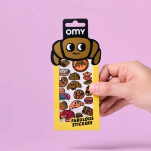 A hand holds a pack of Criossant - Fabulous Stickers, featuring cartoon croissants and pastries on a yellow backing—ideal for pairing with playful sticker earrings—set against a pink background. Gate21.ee